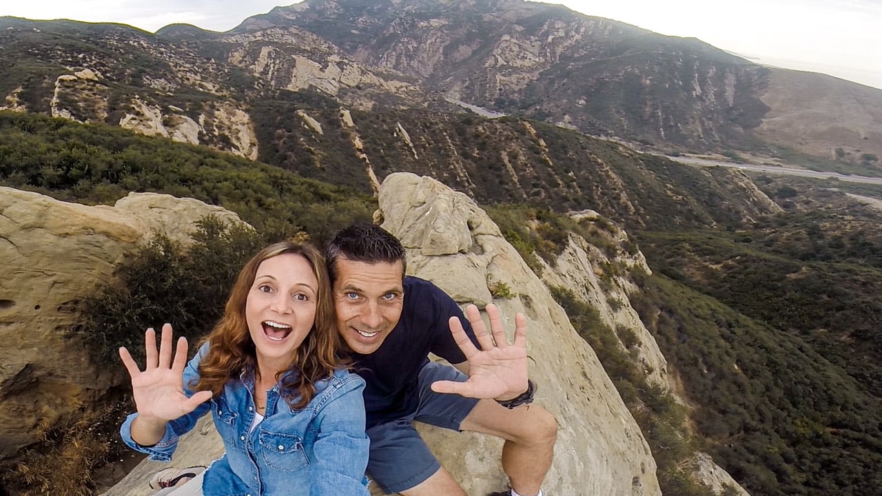 couple sitting on top of a mountain waving
